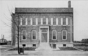 Black and white photograph of the Leland Powers School of the Theater in Boston. The building is a symmetrical, two-story brick structure with a classical design. It features arched windows on the first floor and rectangular windows with shutters on the second floor. A grand entrance with a columned portico and a decorative balcony sits at the center. Ornamental reliefs adorn the top of the facade. A bare tree stands in the foreground, and an industrial landscape is visible in the background.