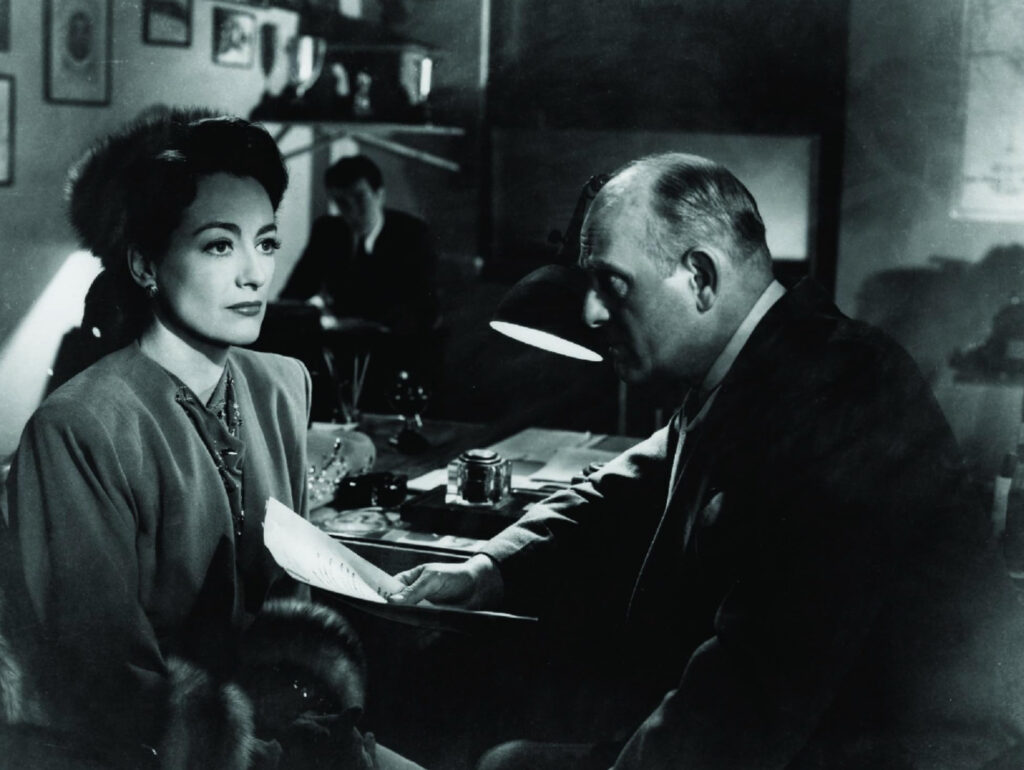 Alt Text:
A black-and-white film still from Mildred Pierce featuring Joan Crawford and Moroni Olsen in a dimly lit office. Crawford, dressed in an elegant suit with a fur stole, sits with a composed expression as Olsen, portraying Inspector Peterson, leans forward and hands her a document. A desk lamp casts dramatic shadows, emphasizing the tense atmosphere. A blurred figure is visible in the background.