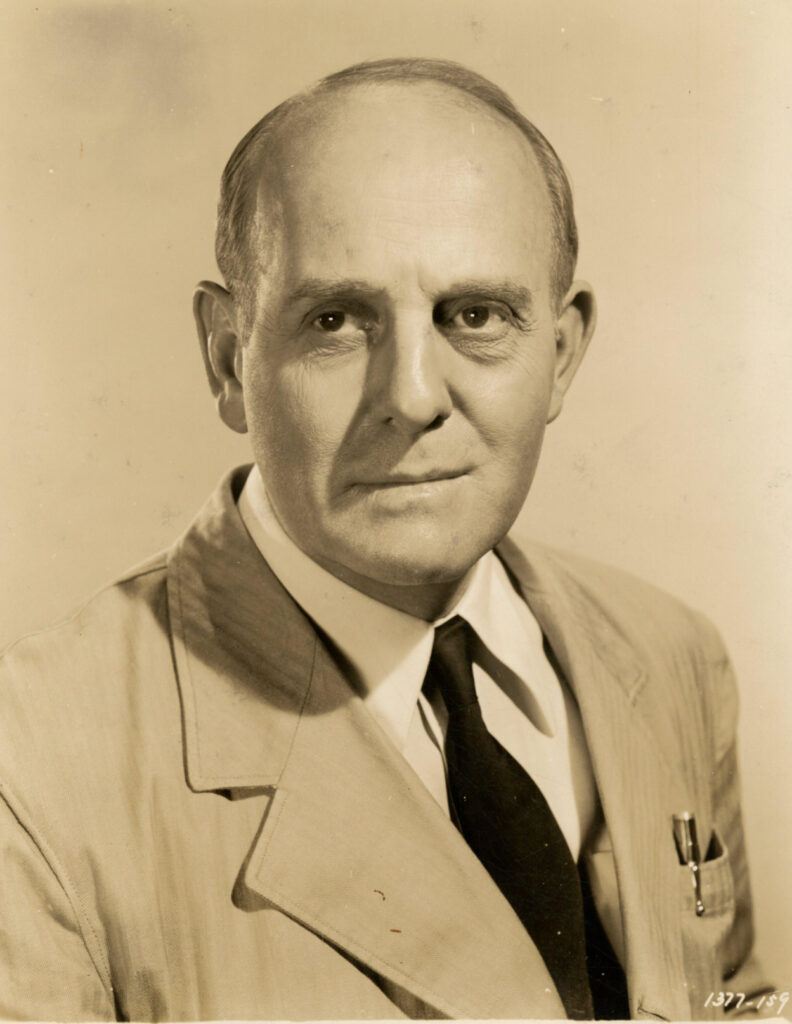 A sepia-toned studio portrait of Moroni Olsen as he appears in the 1947 film The Beginning or the End. He wears a light-colored suit with a dark tie, and a pen is visible in his breast pocket. His expression is serious, with deep-set eyes and a receding hairline. The lighting highlights the contours of his face against a simple background.