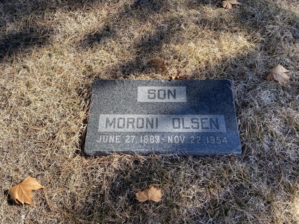 A flat, dark gray granite headstone for Moroni Olsen, set in dry grass with scattered fallen leaves. The inscription reads ‘SON’ at the top, followed by ‘MORONI OLSEN’ and his birth and death dates: ‘JUNE 27, 1889 – NOV 22, 1954.’