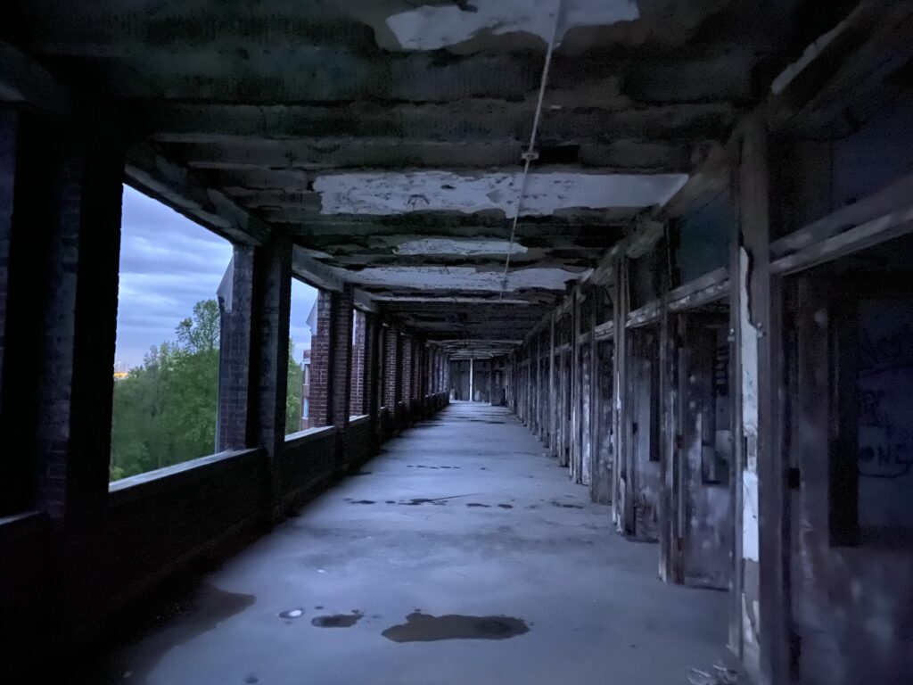 Abandoned hospital corridor at Waverly Hills Sanatorium, peeling ceiling and empty doorways receding into the distance, blue-toned light from open windows on the left