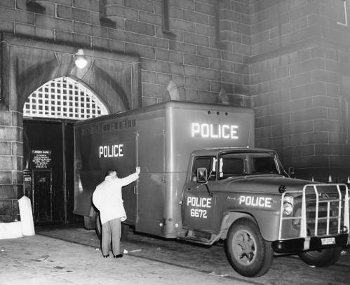 A 1950s Philadelphia Police van backs through the arched south gate entrance of Moyamensing Prison at night, with a figure standing at the rear doors. A sign reading “South Gate, Untried Department” is visible on the left door frame.